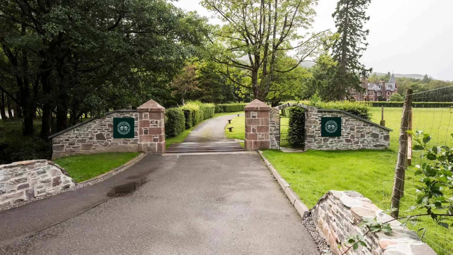 Driven Clay Co. crest signs in forest green on the stone gateposts of a country estate drive, a large house beyond the trees