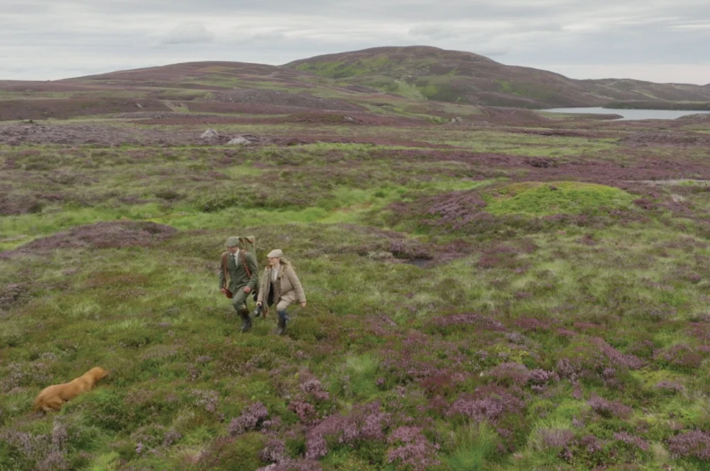 Driven Clay Co. photography: two shooters in tweed crossing purple heather moorland with a gundog, hills and a loch behind