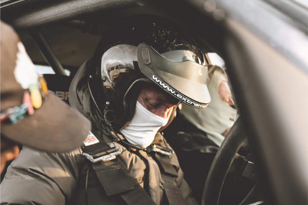 Sportech Motorsport participant in helmet, face covering and harness inside a rally car, with a second occupant beside them