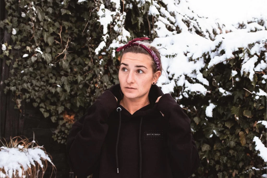 Sportech Motorsport hoodie: small chest logo on black, worn by a woman in a bandana beside snow-covered ivy