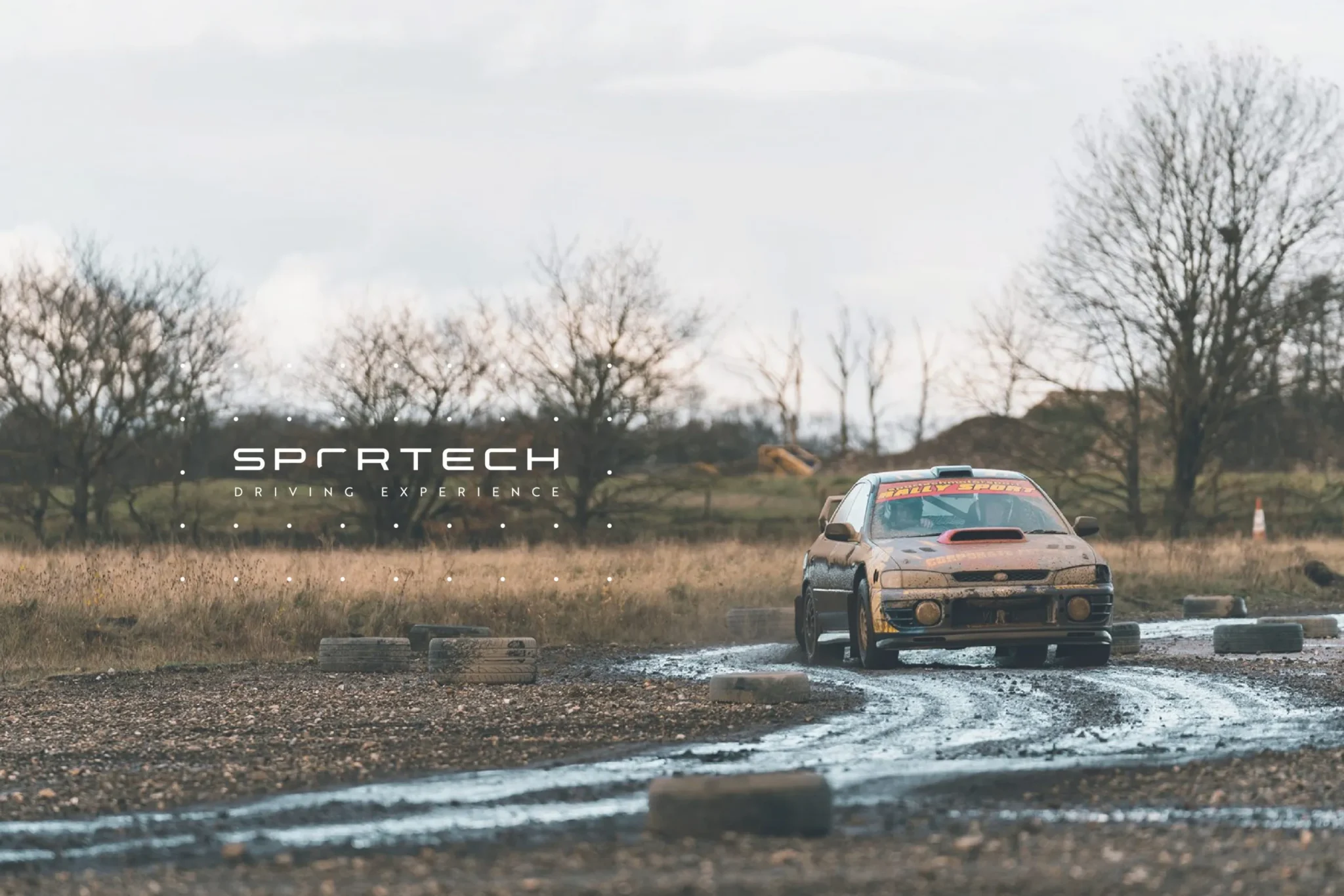 Mud-covered rally car cornering on a wet gravel track past tyre markers, with the Sportech Driving Experience logo