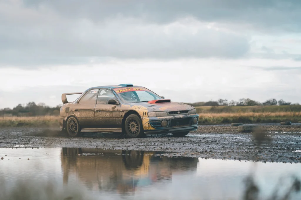 Sportech Motorsport rally car caked in mud on a gravel track, reflected in a large puddle under a cloudy sky