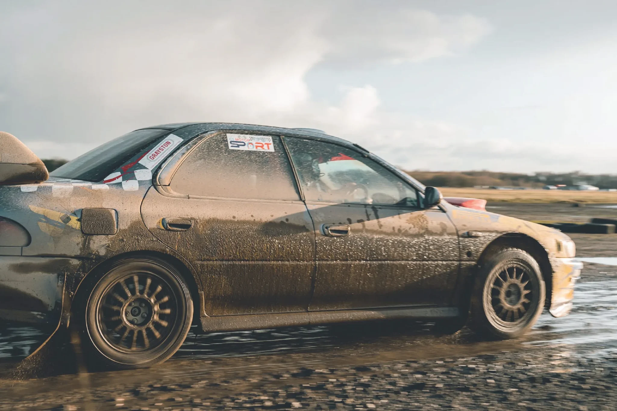 Sportech Motorsport rally car, side-on and splattered with mud, driving through water on a gravel track