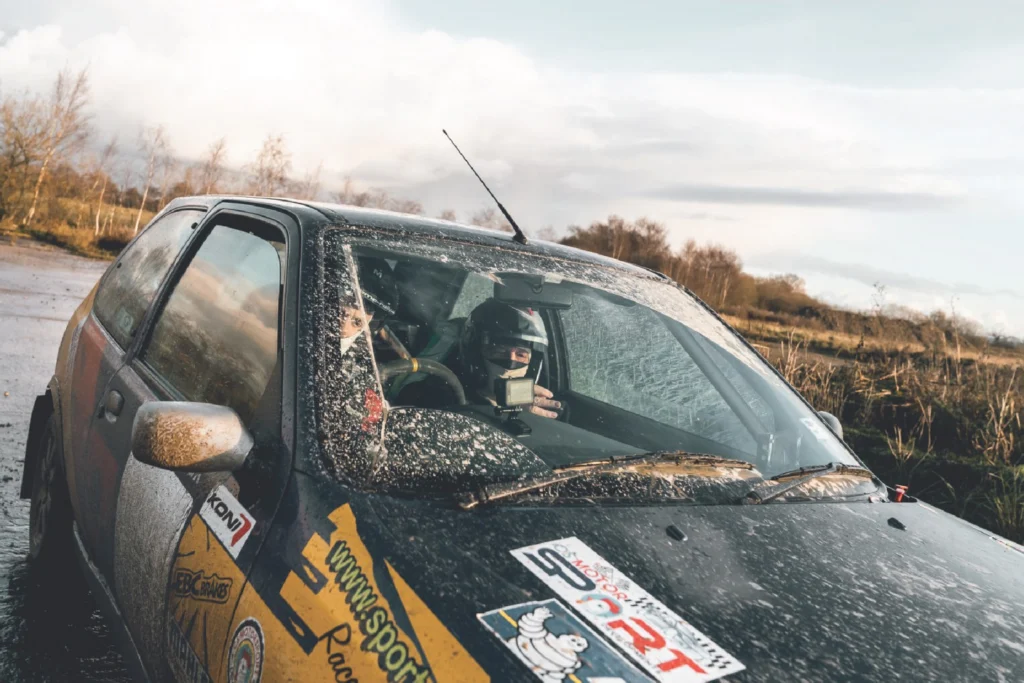 Sportech Motorsport hatchback with sponsor decals, helmeted occupants and a camera seen through its mud-flecked windscreen