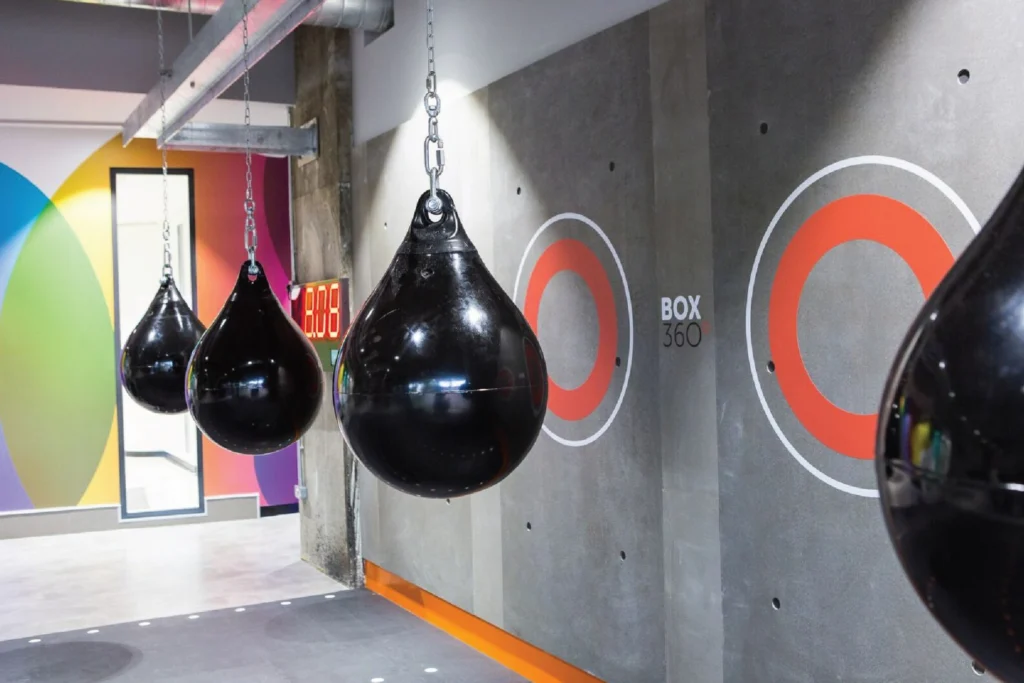 Sweat! Gym boxing area: black water punch bags in front of a concrete wall with orange ring graphics and Box 360 signage