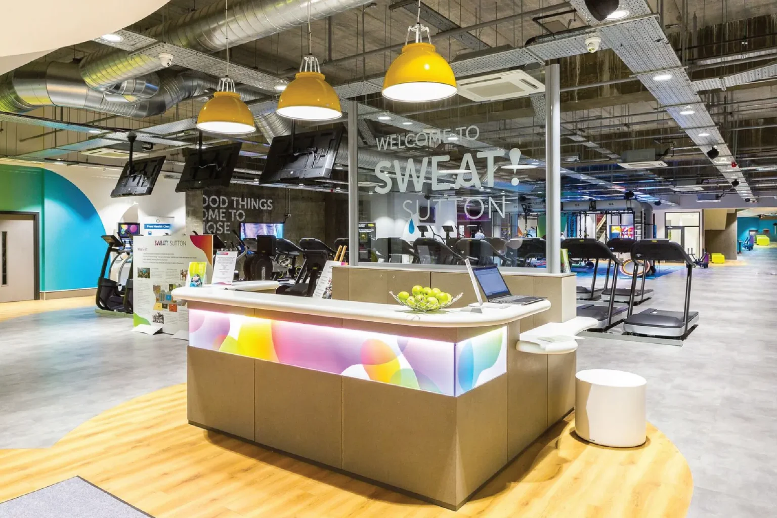 Sweat! Gym reception desk wrapped in colourful bubble graphics, with a Welcome to Sweat! sign on glass and treadmills behind