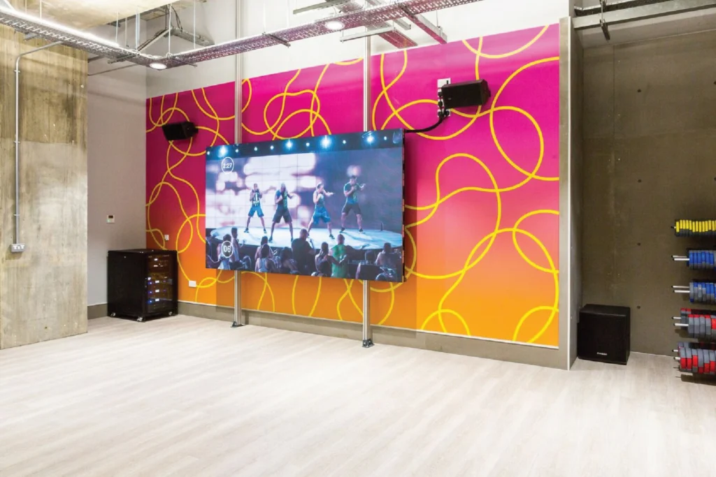 Sweat! Gym studio with a pink-to-orange wall graphic of looping yellow lines around a large screen showing a class