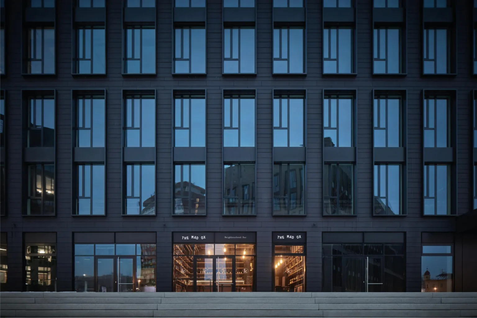 The Mad Ox fascia signs on a glazed ground-floor bar in a dark-clad building at dusk